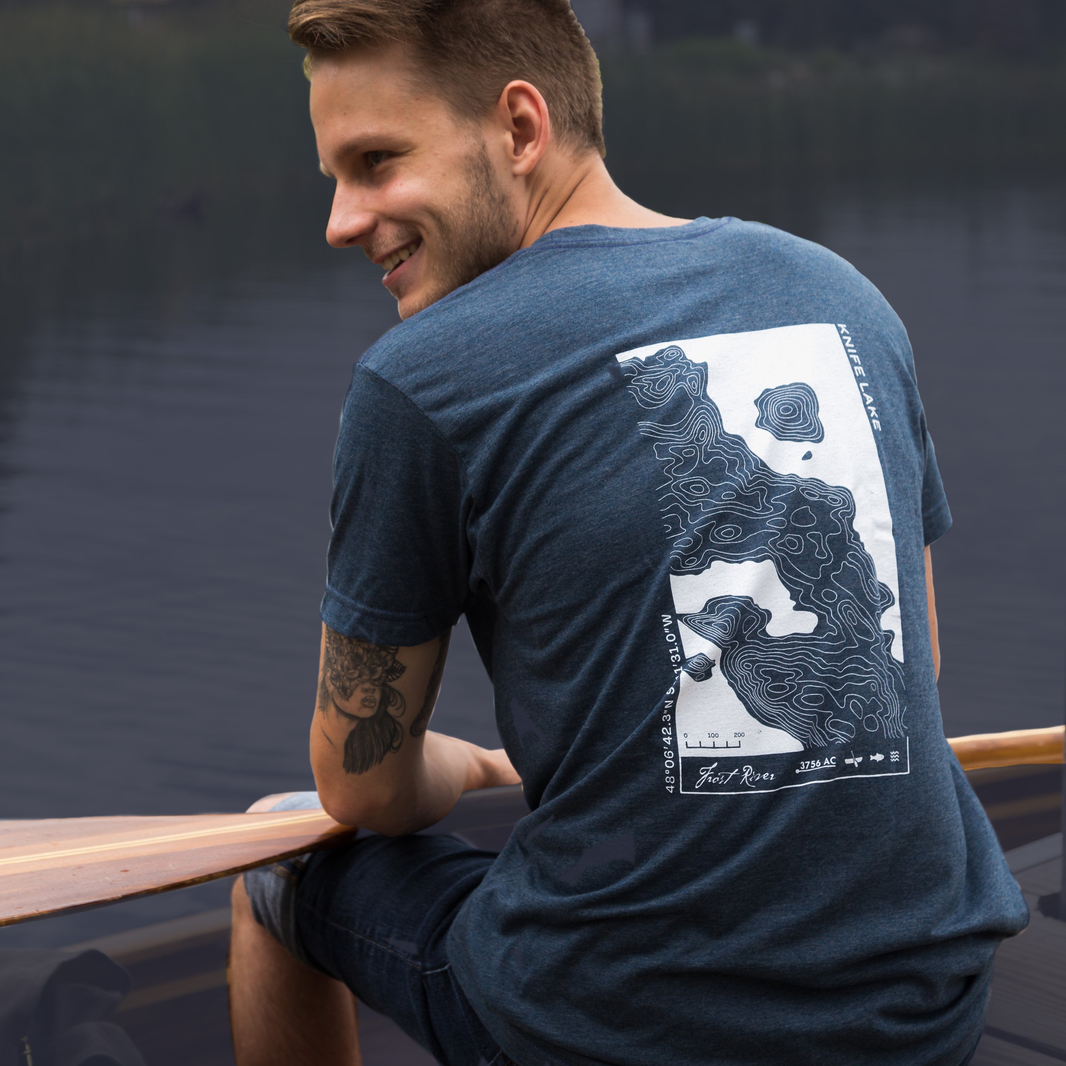Guy sitting on dock holding canoe paddle wearing a graphic T-shirt.